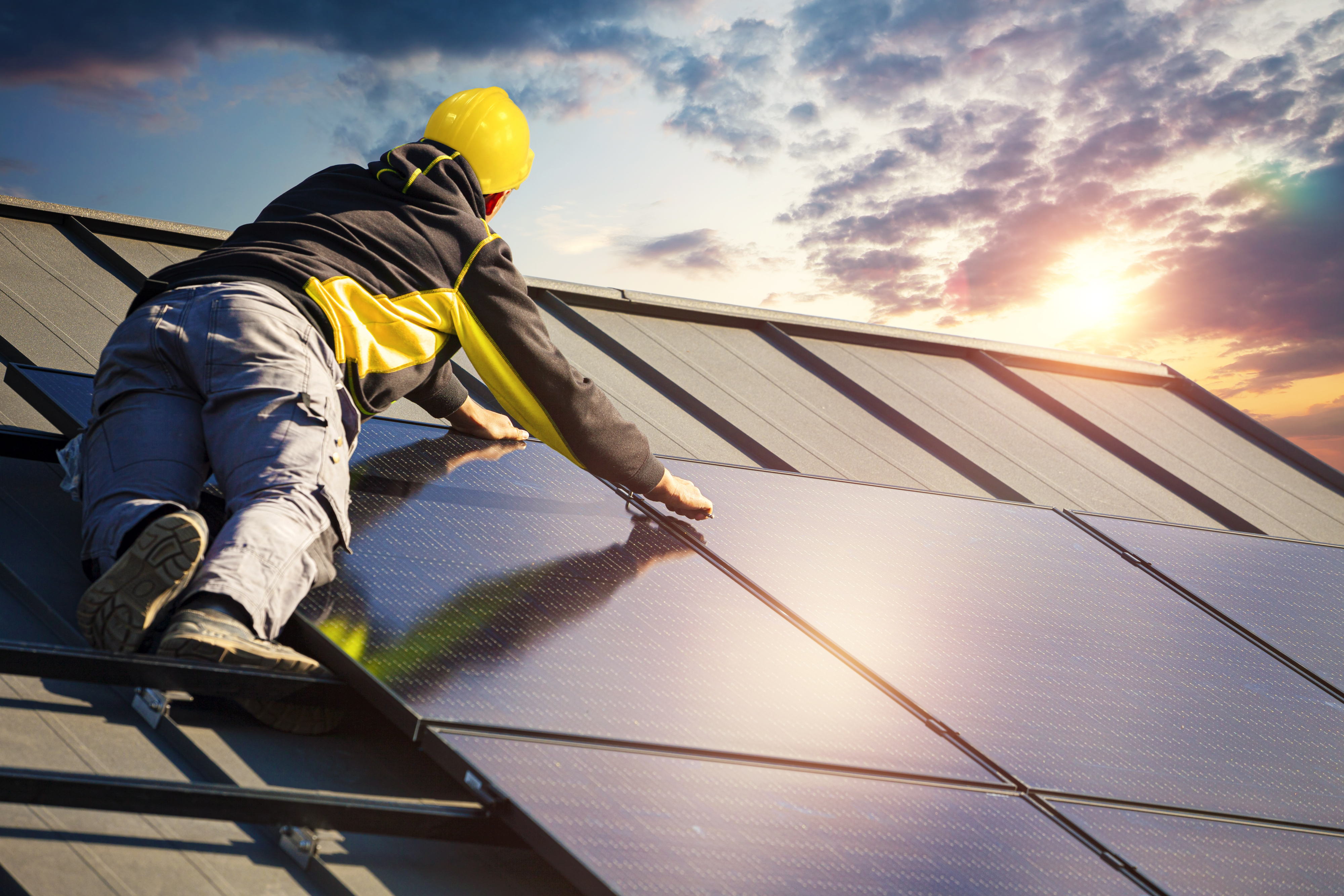 Ein Arbeiter in einem gelben Schutzhelm installiert Solarpanels auf einem Dach bei Sonnenuntergang.