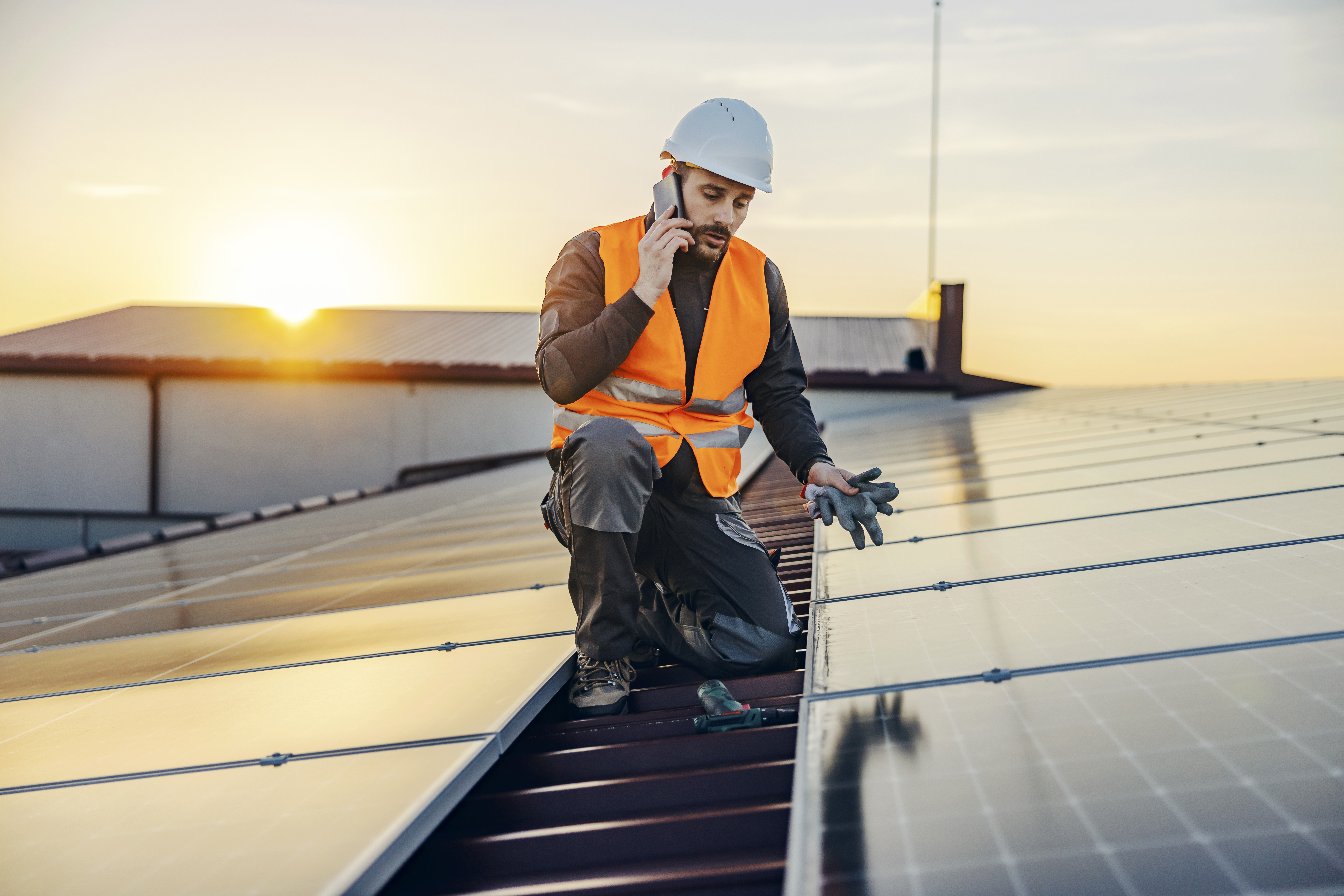 Ein Arbeiter in einer orangenen Warnweste und weissem Schutzhelm kniet auf einem Dach mit Solarpanels und spricht in ein Handy, während die Sonne untergeht.
