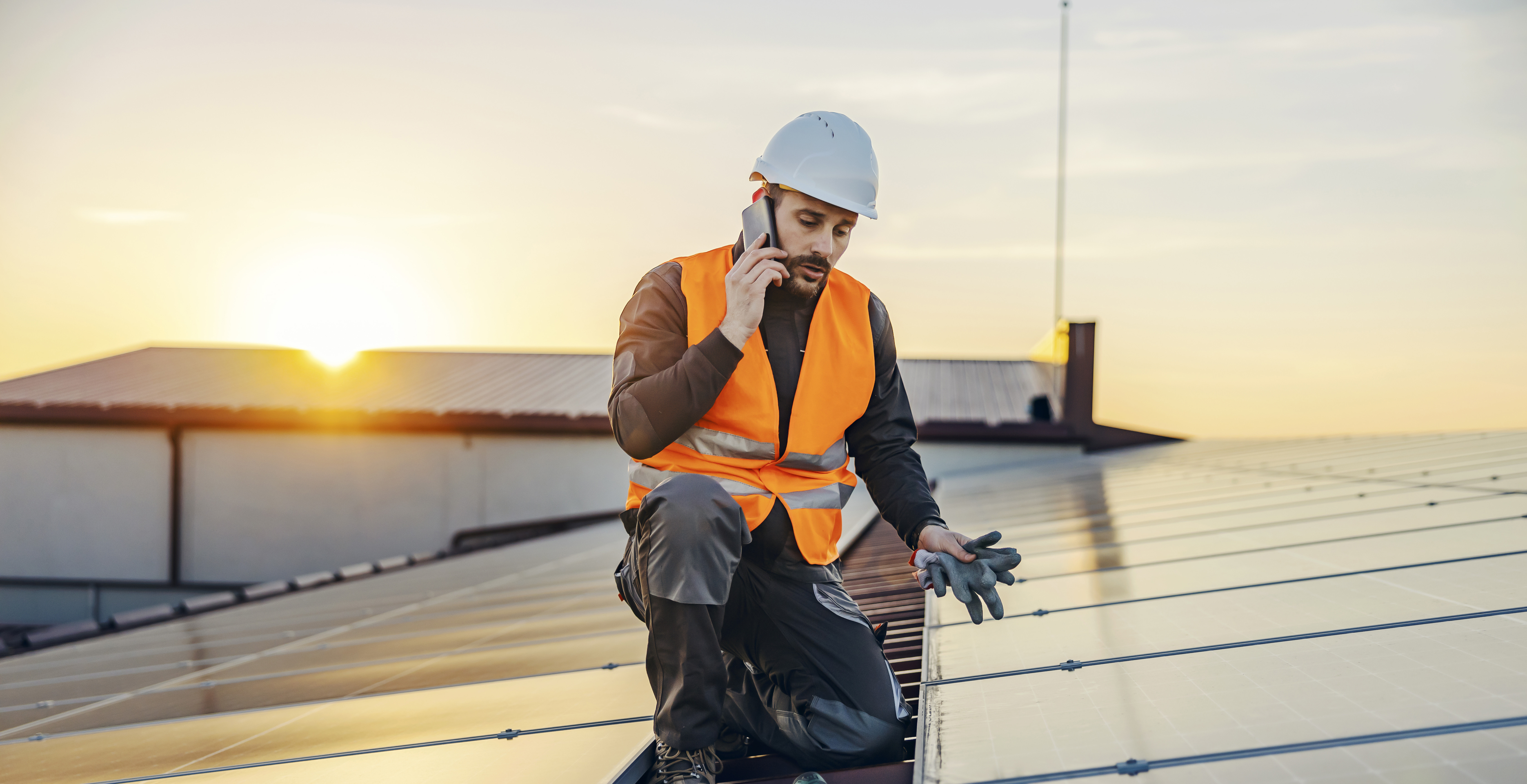 Ein Arbeiter in einer orangenen Warnweste und weissem Schutzhelm kniet auf einem Dach mit Solarpanels und spricht in ein Handy, während die Sonne untergeht.