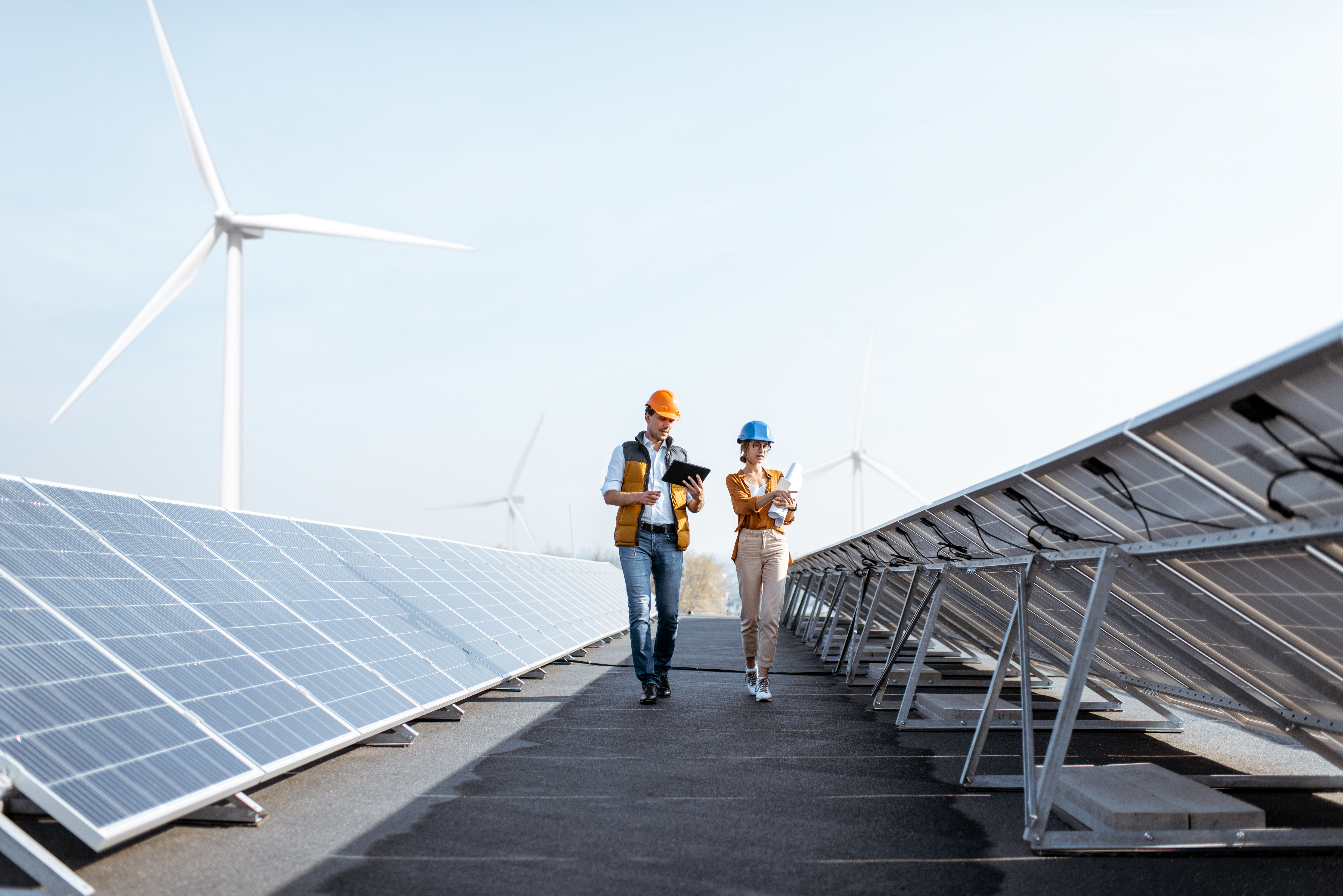 Zwei Ingenieure in Schutzhelmen und Warnwesten inspizieren eine Solaranlage im Freien. Im Hintergrund sind mehrere Windräder zu sehen.