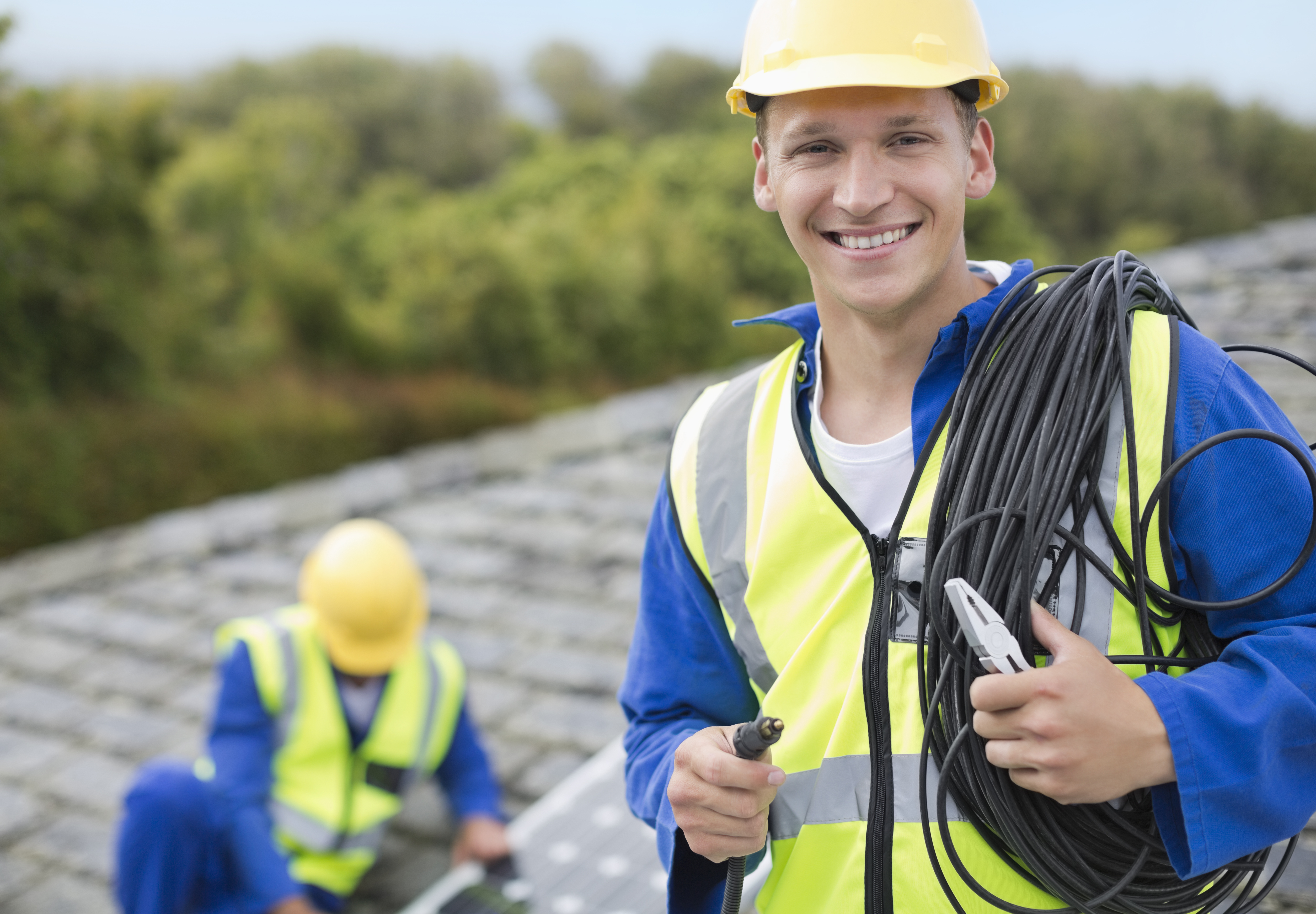 Ein lächelnder Arbeiter in gelbem Schutzhelm und Warnweste hält ein Bündel Kabel und Zangen. Im Hintergrund arbeitet ein weiterer Arbeiter an einem Solarpanel auf einem Dach.