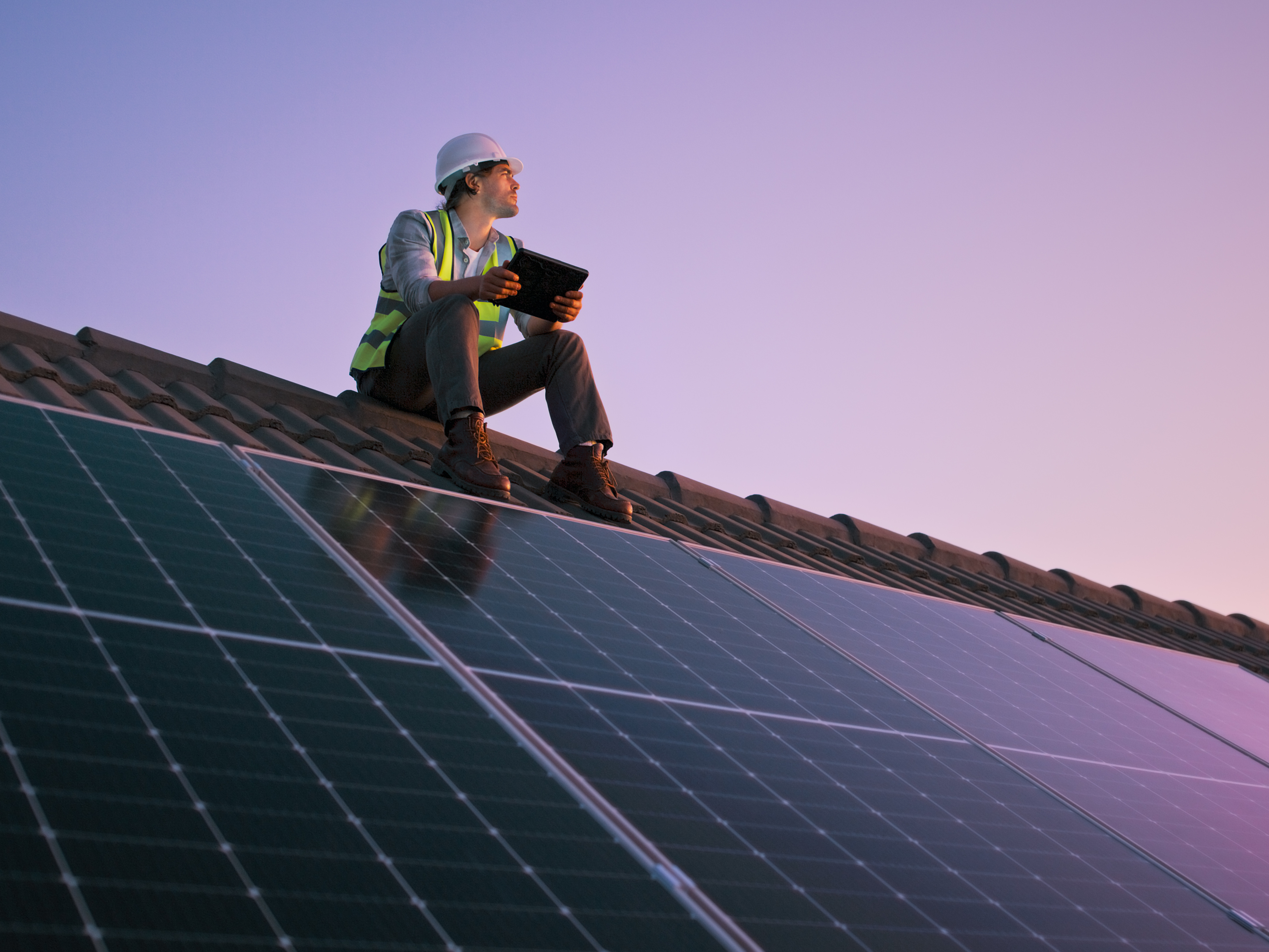 Ein Arbeiter in Schutzhelm und Warnweste sitzt auf einem Dach mit Solarpanels und hält ein Tablet, während er in die Ferne schaut. Der Himmel ist in den Farben des Sonnenuntergangs erleuchtet.