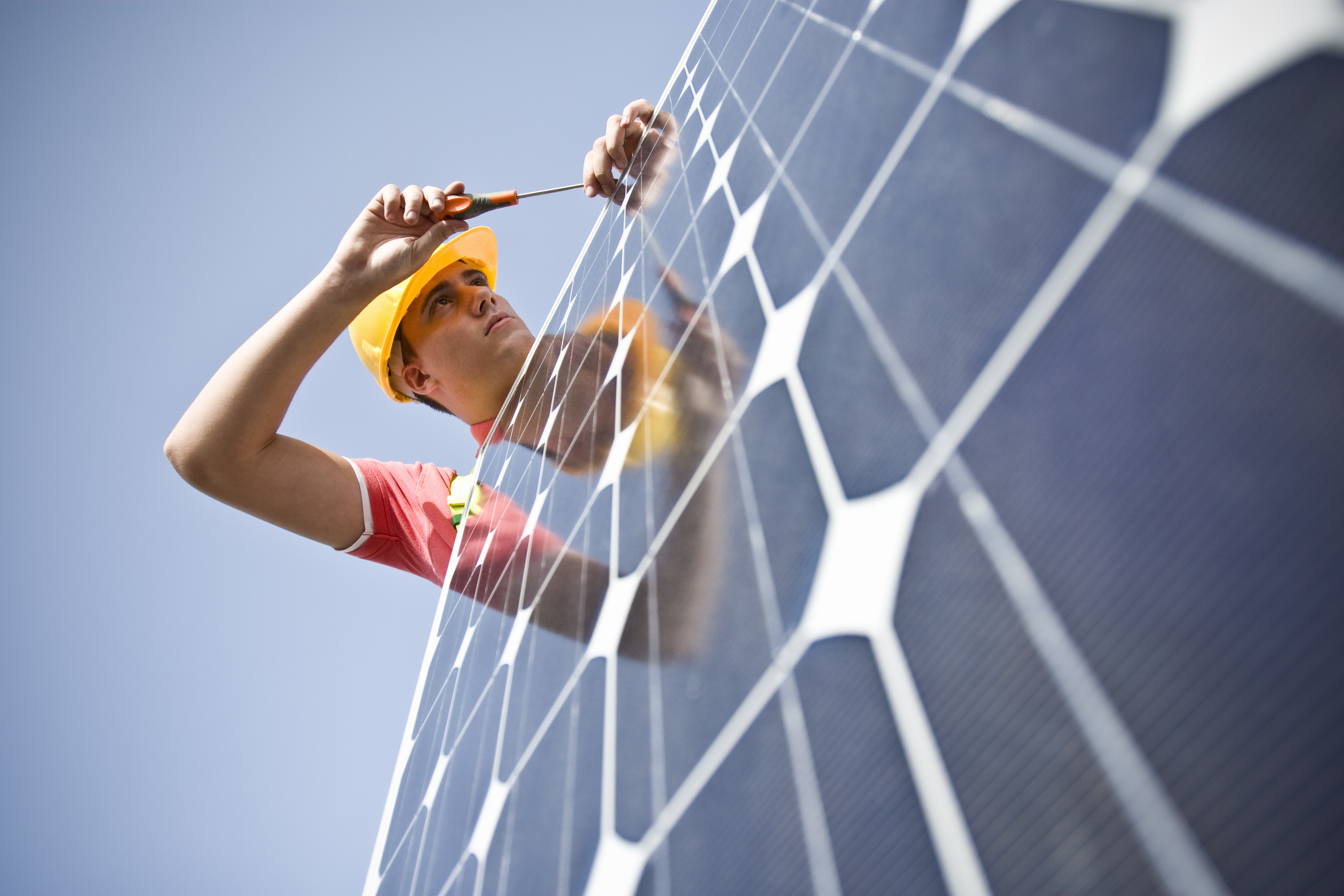 Ein Arbeiter in einem gelben Schutzhelm und rotem Hemd installiert mit einem Schraubenzieher ein Solarpanel unter klarem Himmel.