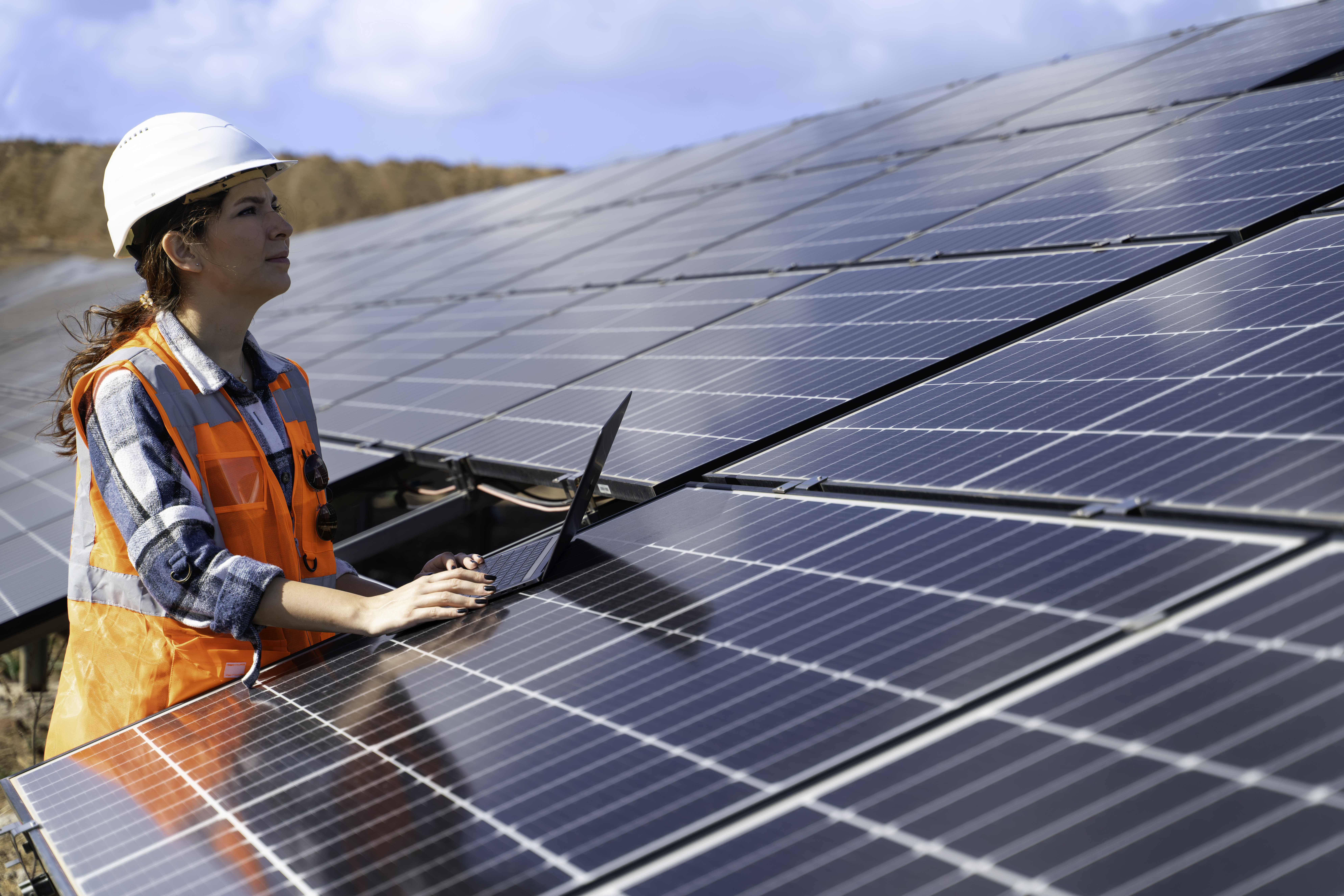 Eine Frau in Schutzhelm und orangener Warnweste arbeitet draussen an einem Laptop und überprüft Solarpanels.