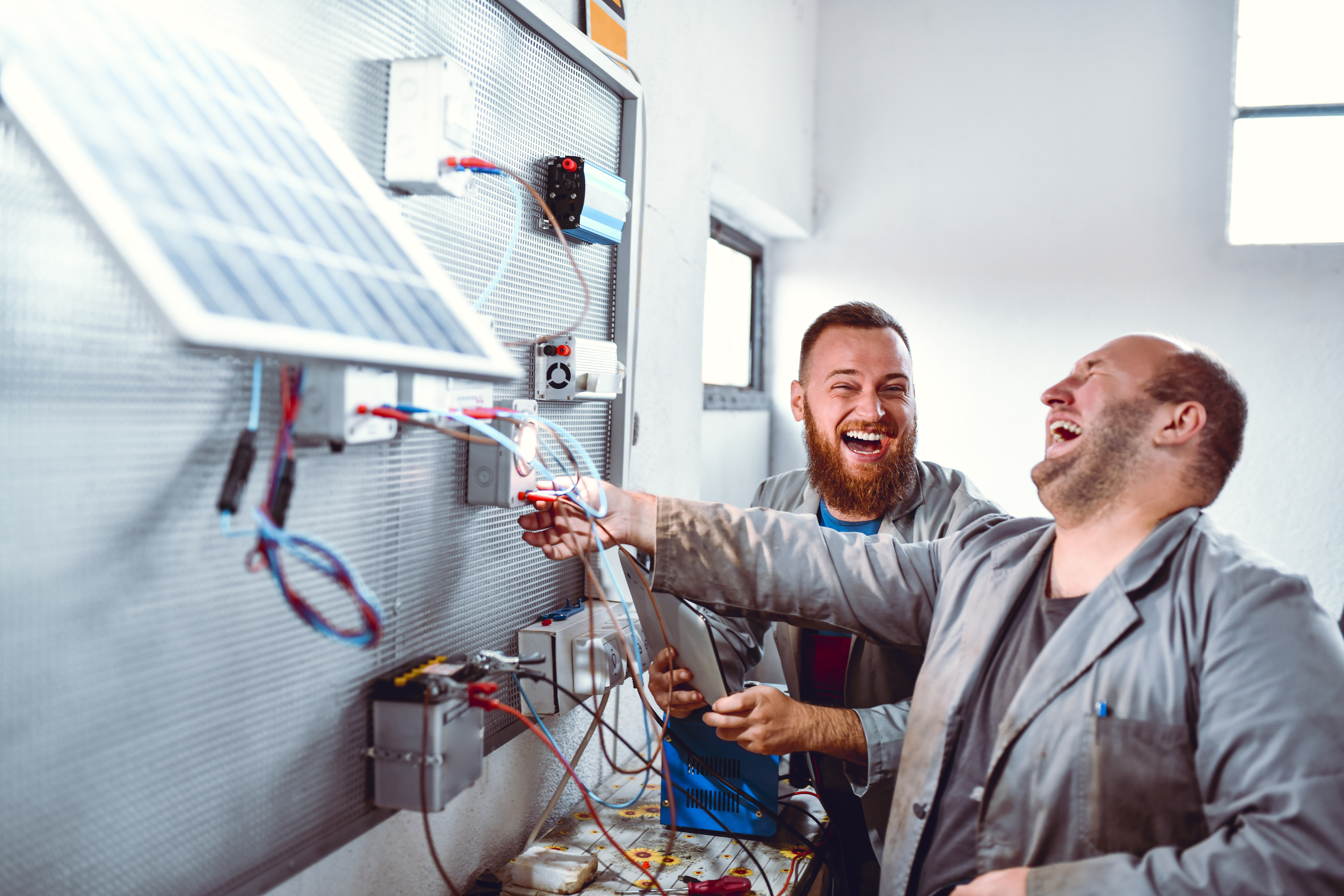 Zwei lachende Männer in grauen Kitteln arbeiten an einem Solarpanel-Installationsprojekt in einem Innenraum. Einer der Männer hält ein Werkzeug und justiert Kabel.
