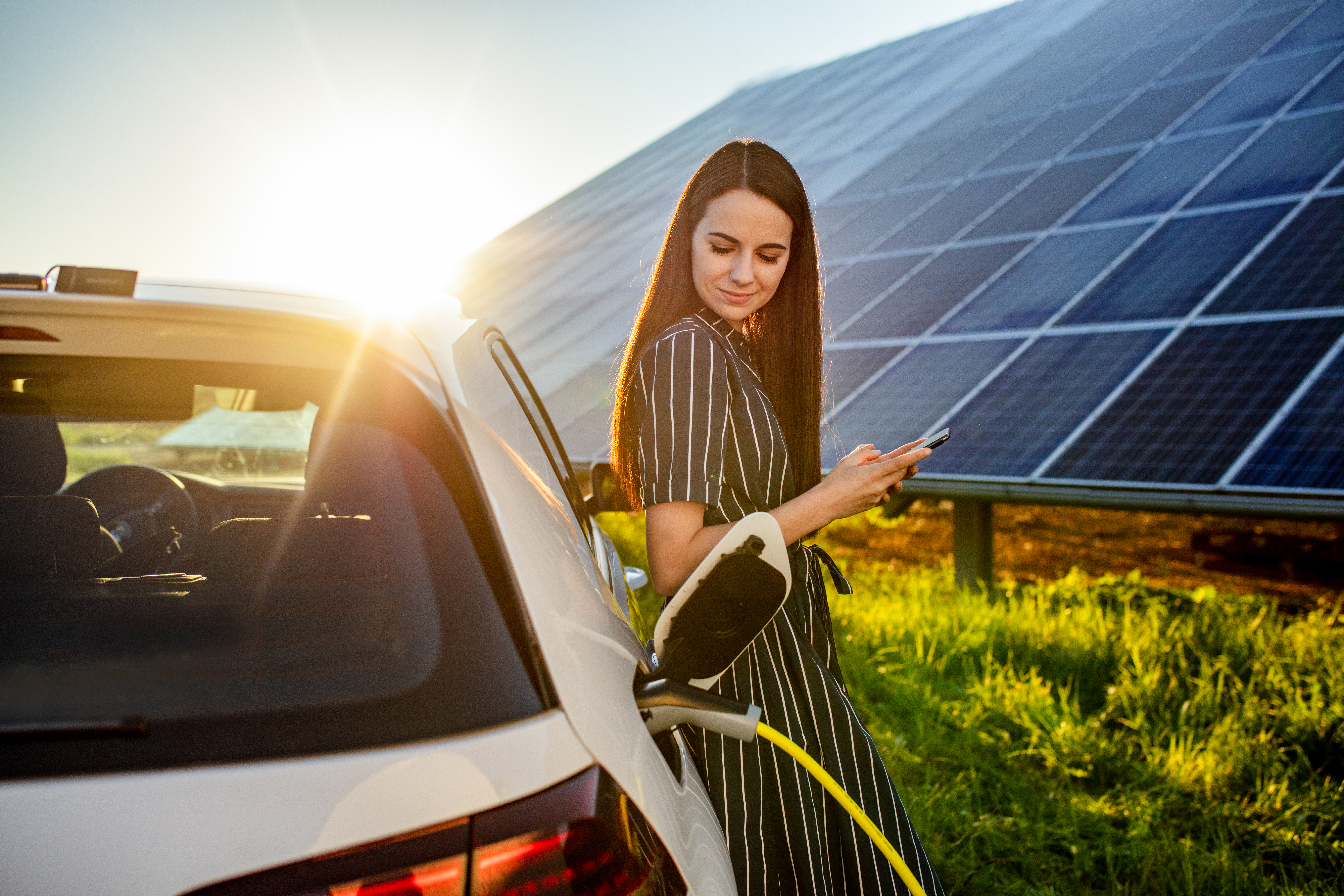 Eine Frau in einem gestreiften Kleid lädt ihr Elektroauto neben einem Solarpanel-Feld auf und benutzt dabei ihr Smartphone. Die Sonne geht im Hintergrund unter.
